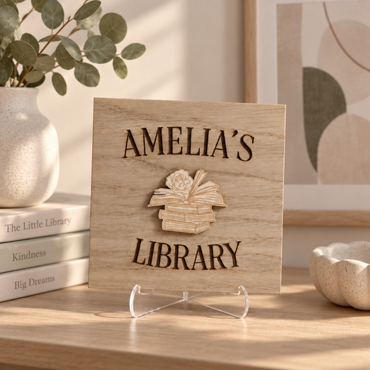 A personalized wooden sign reading 'AMELIA'S LIBRARY' with a raised book stack design in the center, displayed on a clear acrylic stand on a wooden table in a styled interior setting.