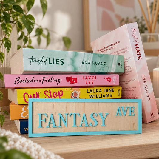 A decorative wooden sign reading 'FANTASY AVE' in raised turquoise letters with a turquoise frame, styled on a wooden shelf in front of a stack of colorful books including 'Twisted Lies', 'Booked on a Feeling', 'Our Stop', and 'Fantasy Hate'.