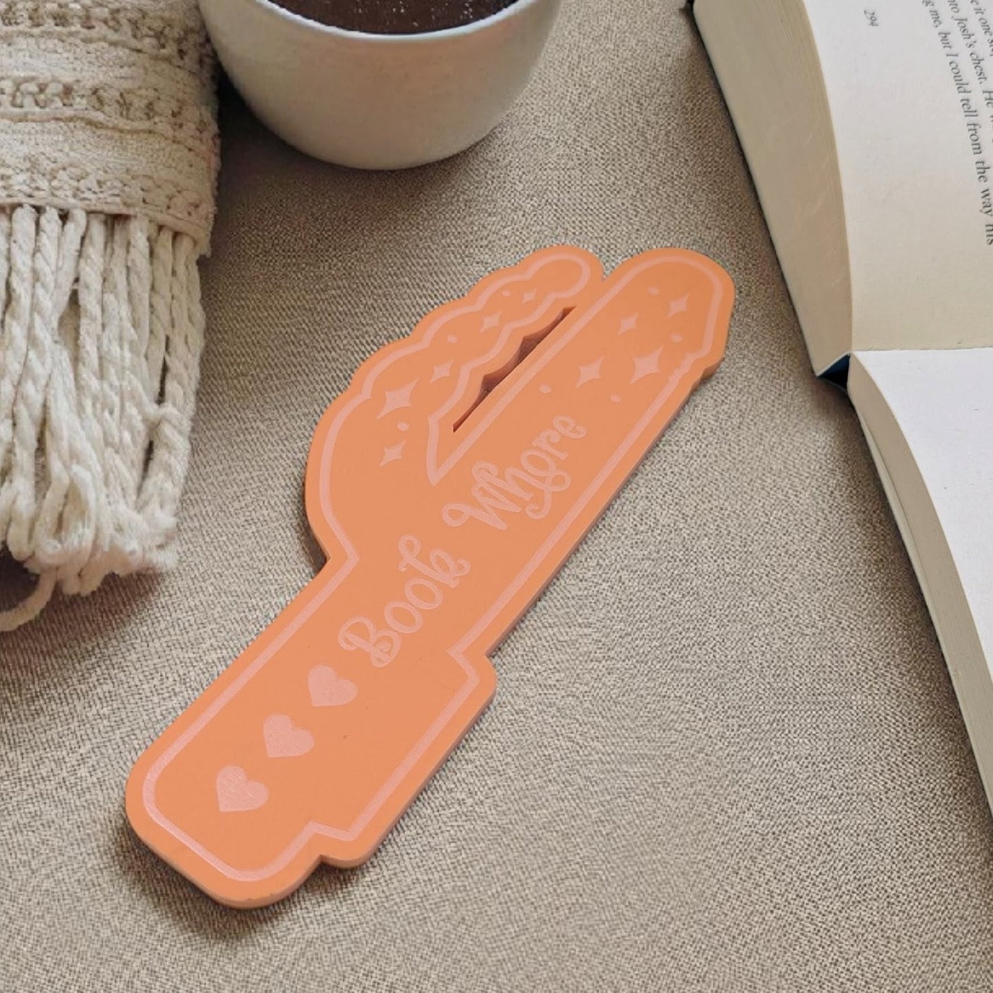 Orange vibrator bookmark with 'Book Whore' text on a textured surface with a cup and book.