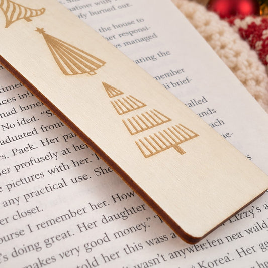 A close-up of a light natural wood bookmark with laser-engraved Christmas tree designs, resting on a book page with a cozy, festive background.