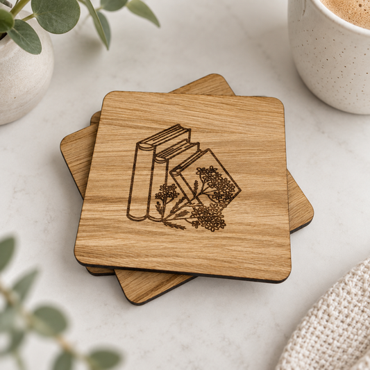 A stack of square wooden coasters with a laser-engraved design of books and herbs, styled on a light marble surface with a ceramic cup and greenery.