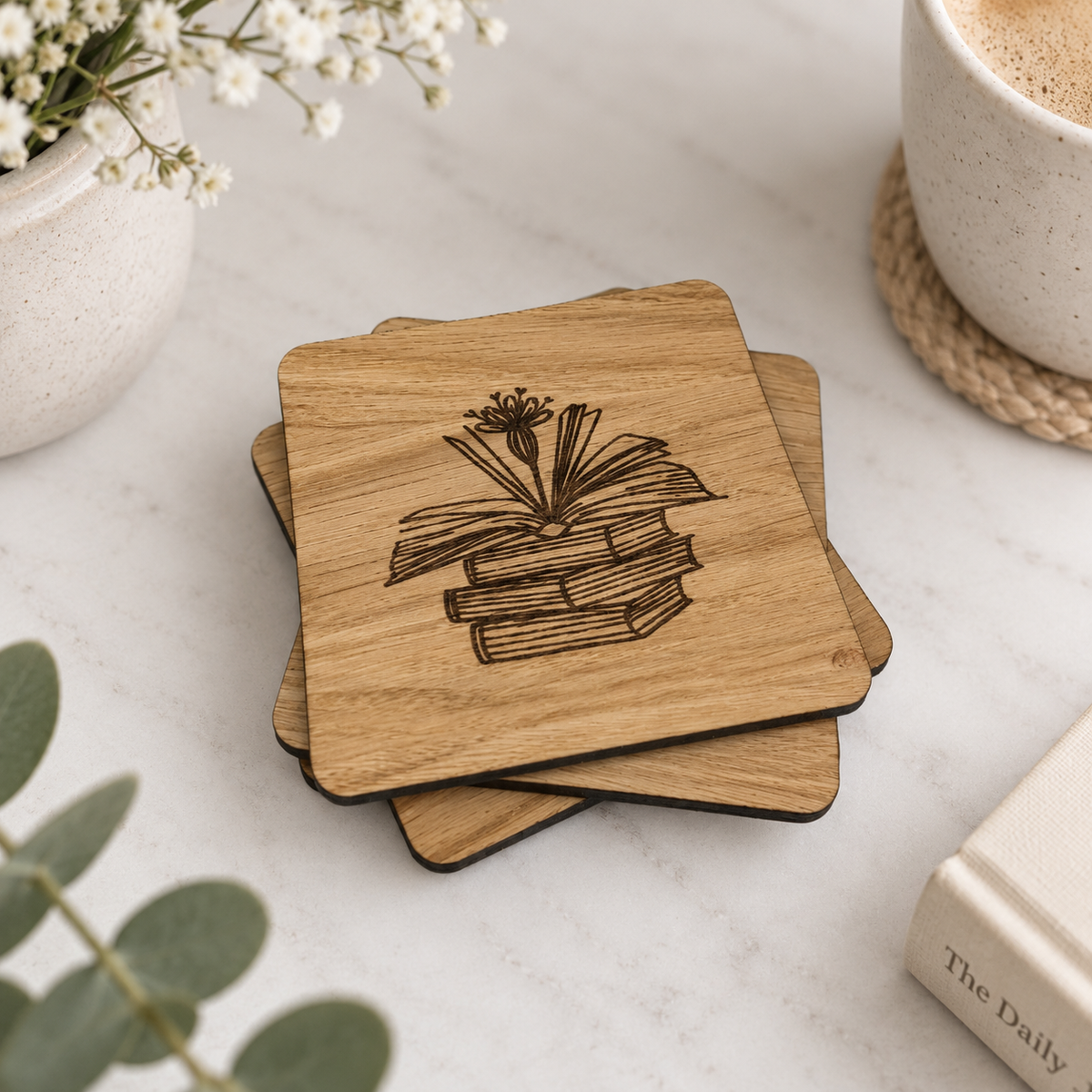 A stack of square wooden coasters with rounded corners, each engraved with a book-and-flower illustration, displayed on a light marble surface with coffee cups and flowers as props.