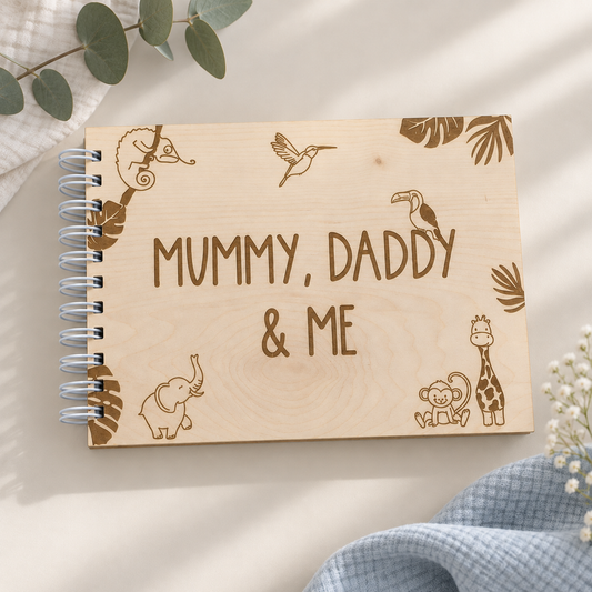A personalized wooden spiral notebook with engraved animal illustrations and the text 'MUMMY, DADDY & ME' on the front cover, styled on a light beige surface with soft fabric and greenery props.