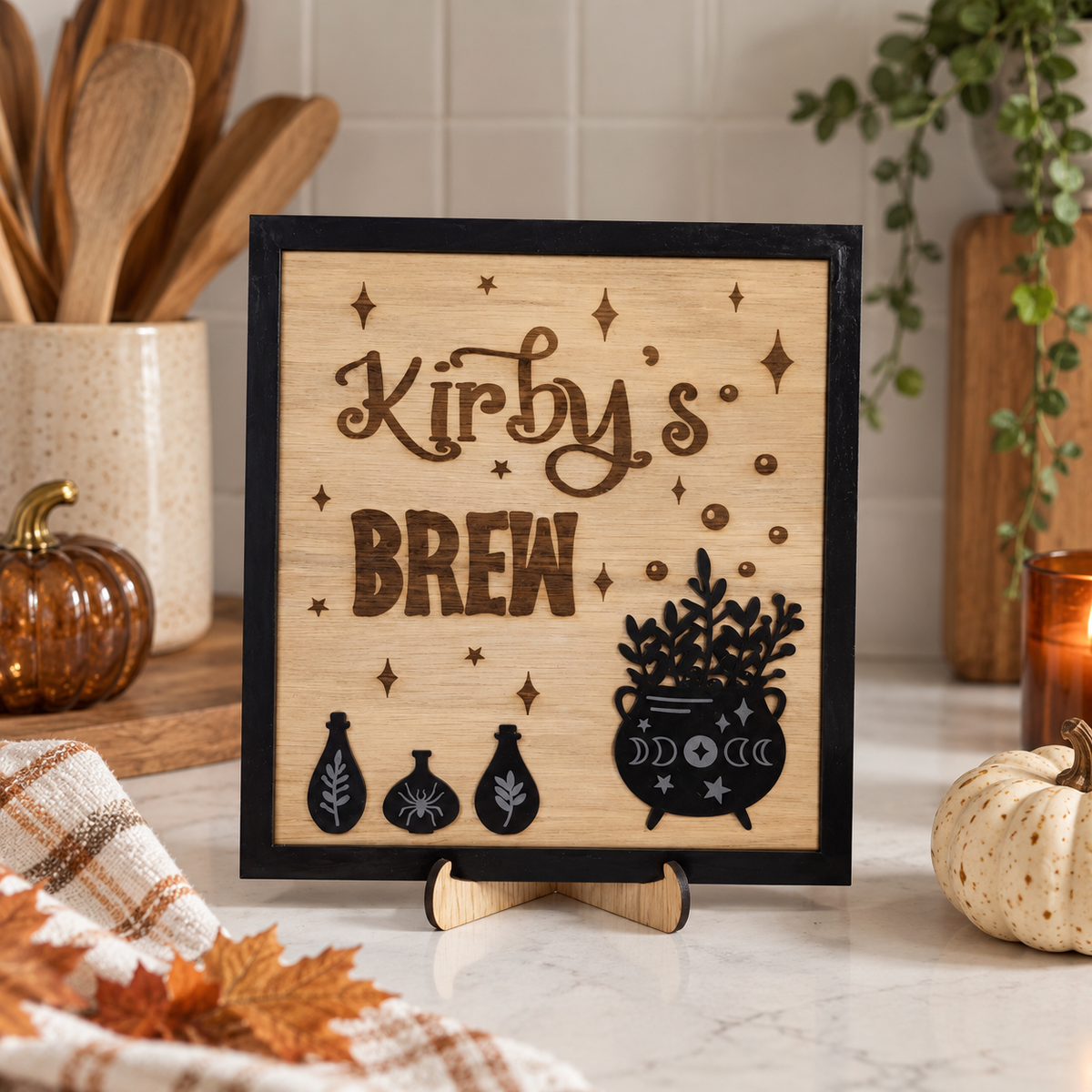 A square wooden sign with a black frame and stand, featuring the engraved text 'Kirby's BREW' with black cauldron, potion bottles, and star motifs on a light wood background. The sign is styled on a marble countertop with autumn-themed props including a small pumpkin, a candle, and wooden utensils.