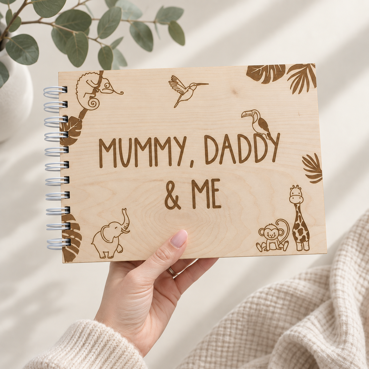 A hand holds a spiral-bound wooden notebook with laser-engraved safari animal illustrations and the text 'MUMMY, DADDY & ME'. The background is a soft beige fabric with a eucalyptus branch in the corner.