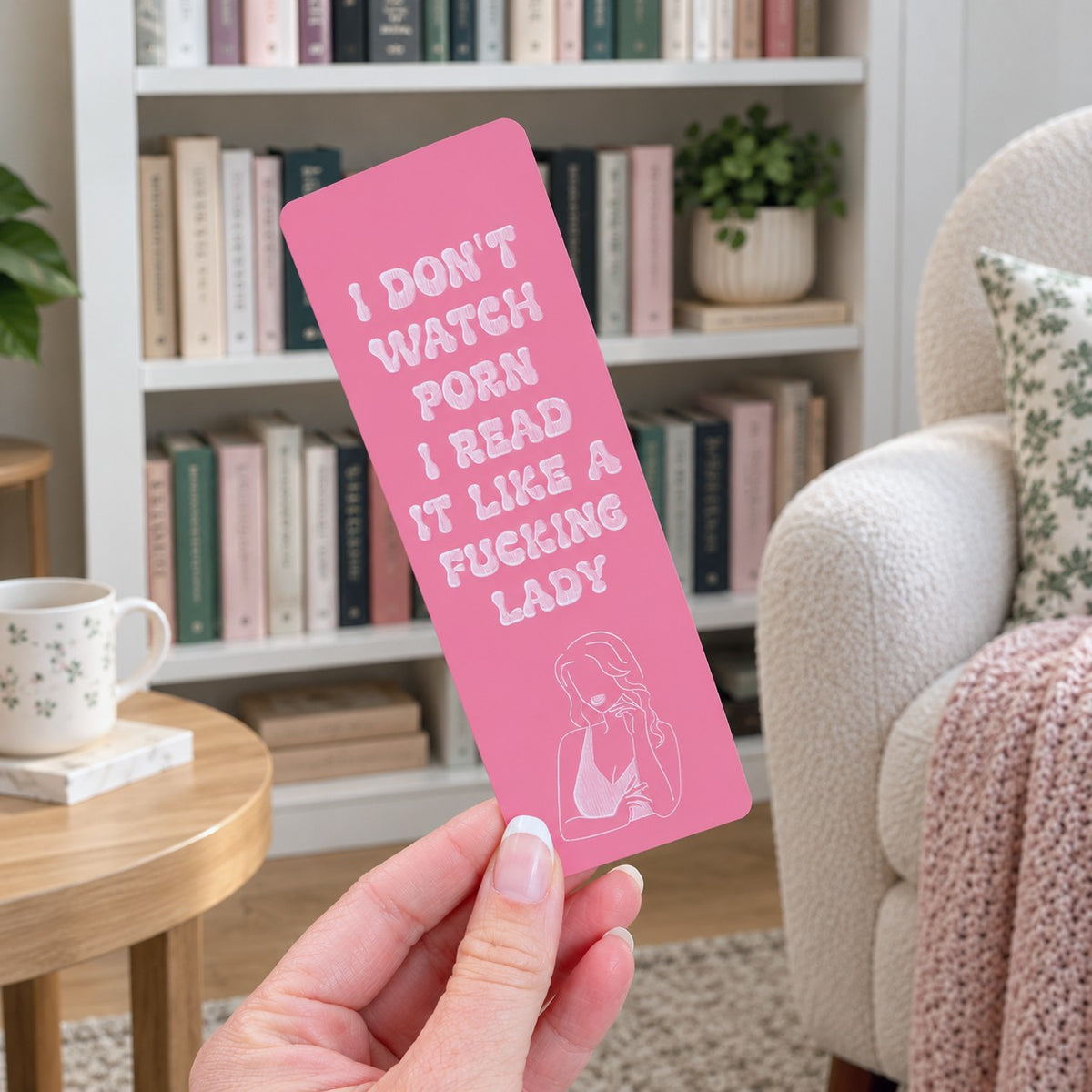 A hand holds a pink rectangular bookmark or card with white text reading 'I DON'T WATCH PORN I READ IT LIKE A FUCKING LADY' and a small line drawing near the bottom, set against a cozy living room background with a bookshelf, chair, and mug.