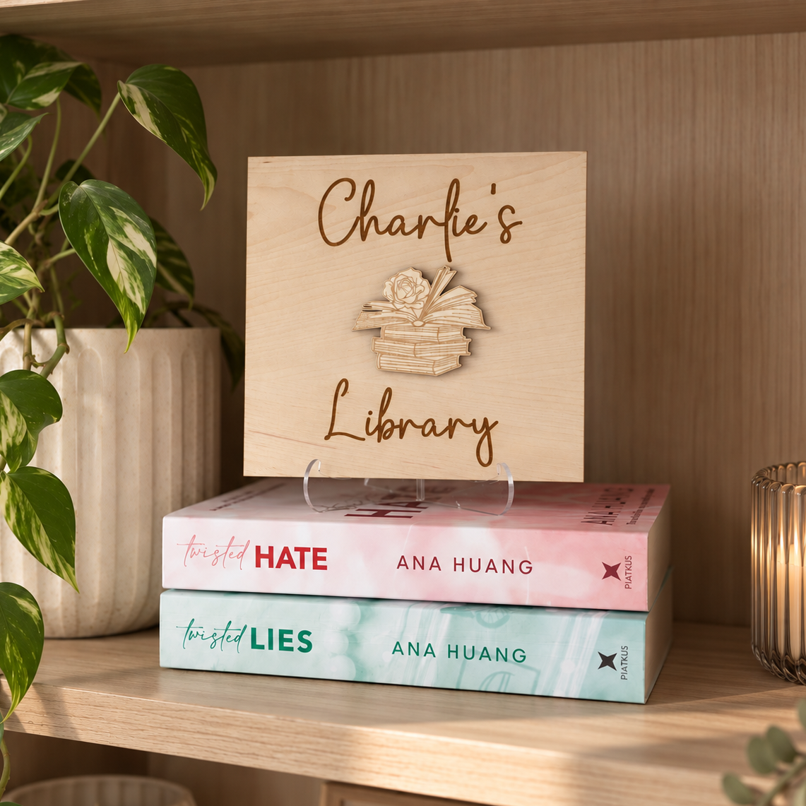 A personalized wooden sign reading 'Charlie's Library' with an engraved book illustration, displayed on a wooden shelf above a stack of three books by Ana Huang.