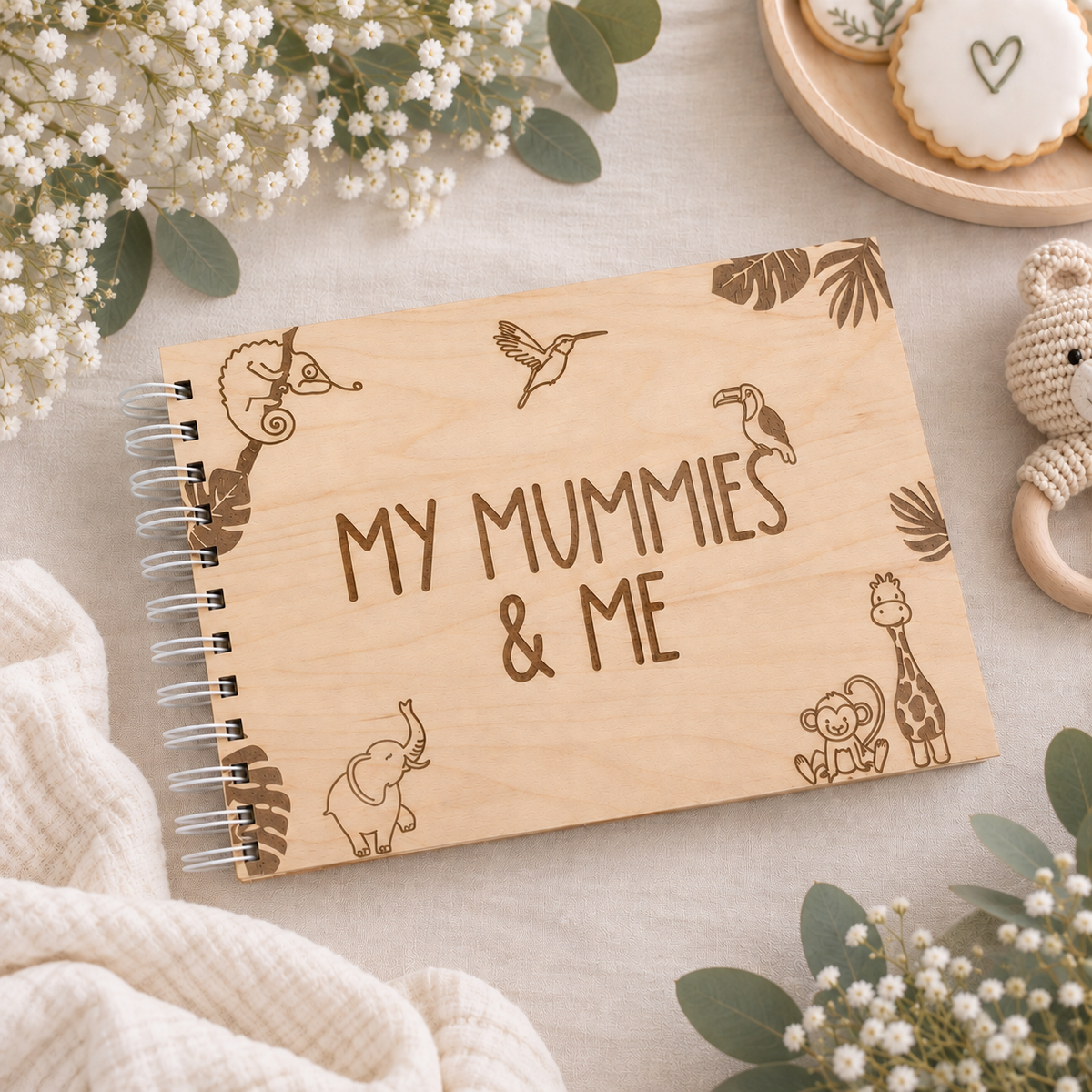 A personalized wooden spiral notebook with 'MY MUMMIES & ME' engraved in the center, surrounded by safari animal illustrations. The notebook is styled in a flat-lay setting with baby's breath flowers, a knitted blanket, and a wooden toy.