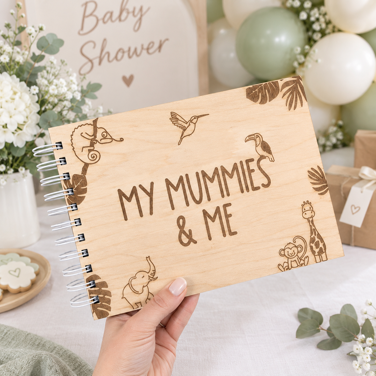 A hand holds a spiral-bound wooden notebook with laser-engraved animal illustrations and the text 'MY MUMMIES & ME'. The background shows a styled baby shower setting with balloons, flowers, and gift boxes.