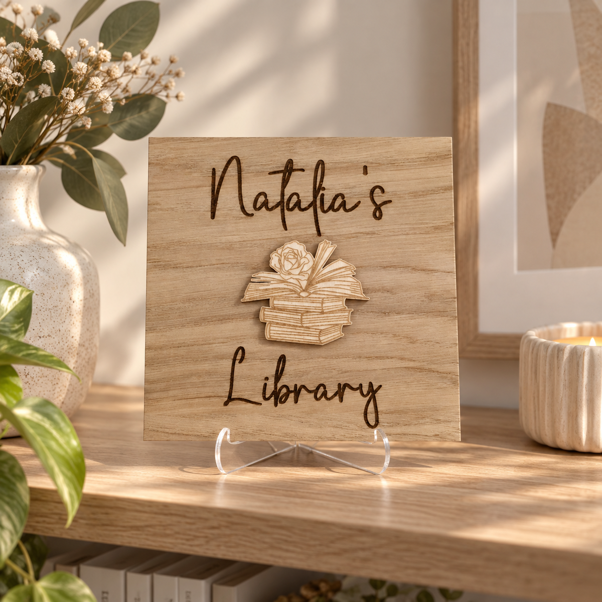 A square wooden sign reading "Natalia's Library" with a 3D-effect book stack design in the center, displayed on a wooden shelf in a styled home setting with a candle, vase, and framed art.