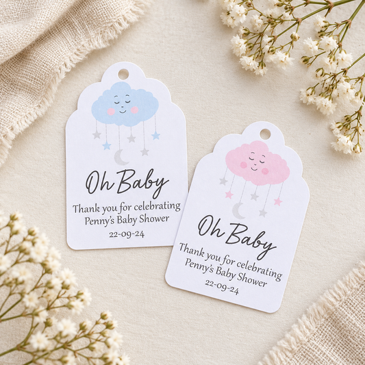 Two white baby shower favor tags with cute cloud illustrations, styled on a beige fabric background with dried white flowers.