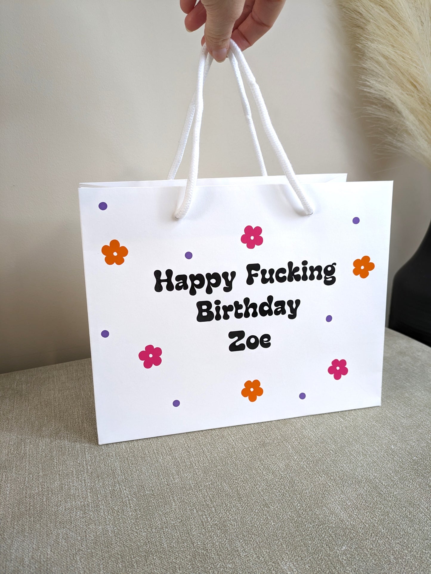 a white gift bag with a rope handle. Each bag has bright pink and orange daisy flowers and the taboo wording.