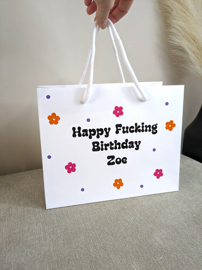 a white gift bag with a rope handle. Each bag has bright pink and orange daisy flowers and the taboo wording.