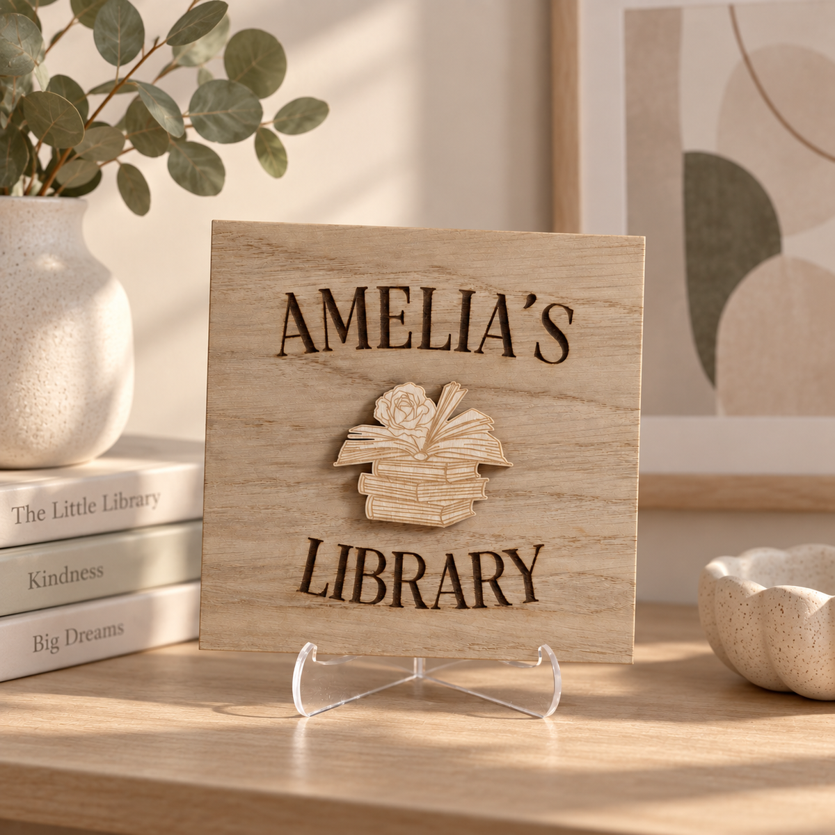 A personalized wooden sign reading 'AMELIA'S LIBRARY' with a raised book stack design in the center, displayed on a clear acrylic stand on a wooden table in a styled interior setting.