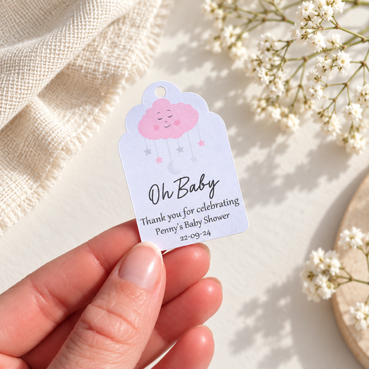 A hand holds a personalized baby shower favor tag shaped like a cloud, printed with a pink smiling cloud, stars, and the text 'Oh Baby Thank you for celebrating Penny's Baby Shower 22.09.