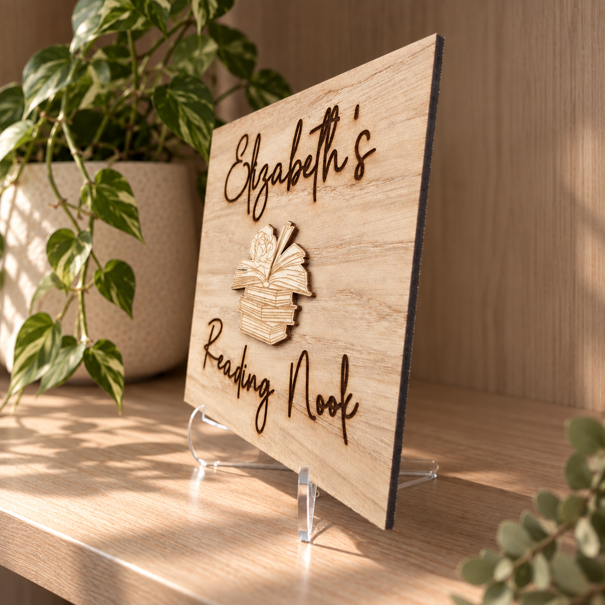 A personalized wooden sign reading 'Elizabeth's Reading Nook' with a small book icon, displayed on a clear acrylic stand on a wooden shelf with houseplants in the background.