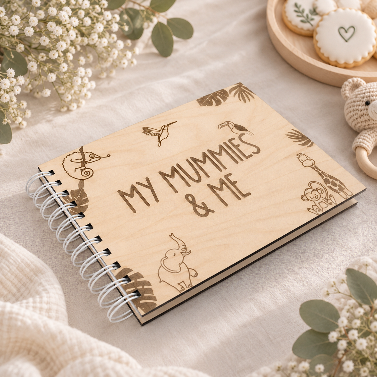 A wooden spiral notebook with 'MY MUMMIES & ME' engraved on the cover, styled on a beige fabric surface with baby props including dried flowers, cookies, and a teddy bear.