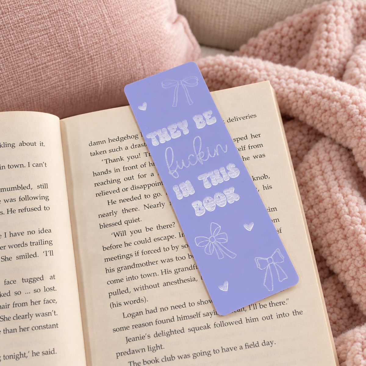 A lavender purple paper bookmark with white line art illustrations and the text 'THEY BE fuckin' IN THIS BOOK' resting on an open book, set against a cozy pink knitted blanket background.