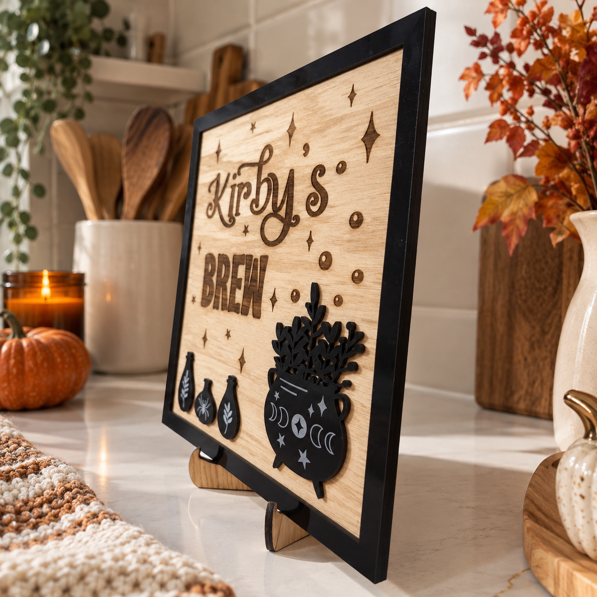 A framed wooden sign with the words 'Kirby's BREW' and black occult-themed cutout decorations, displayed on a countertop in a styled kitchen setting.