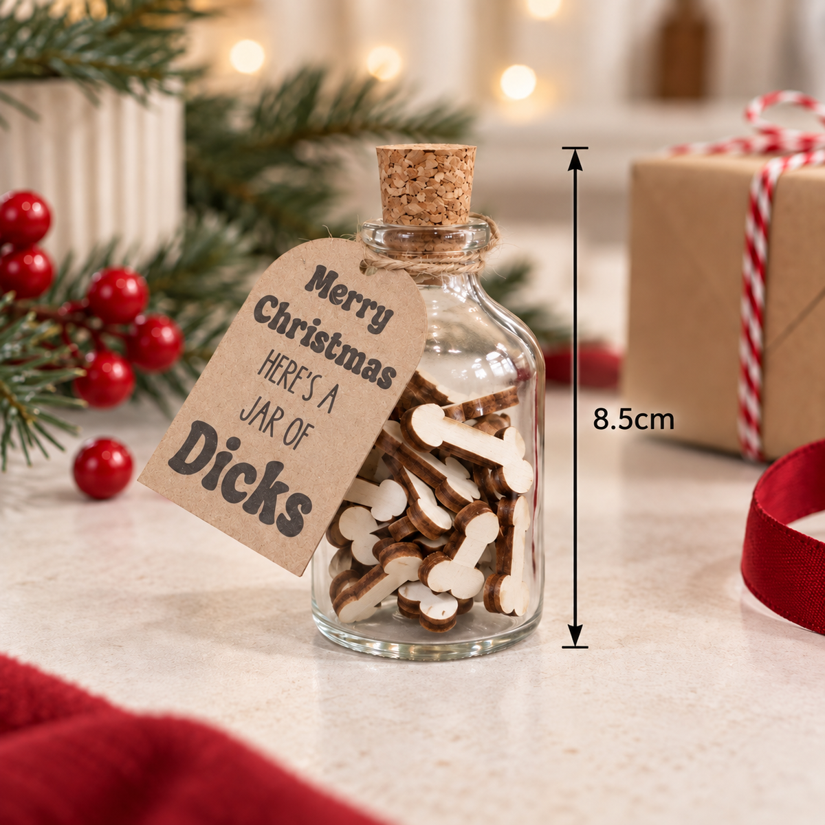 A clear glass bottle with a cork stopper, filled with small PENIS shaped WOODEN PARTS, sits on a light countertop. A kraft paper tag reads “Merry Christmas Here’s a Jar of Dicks.