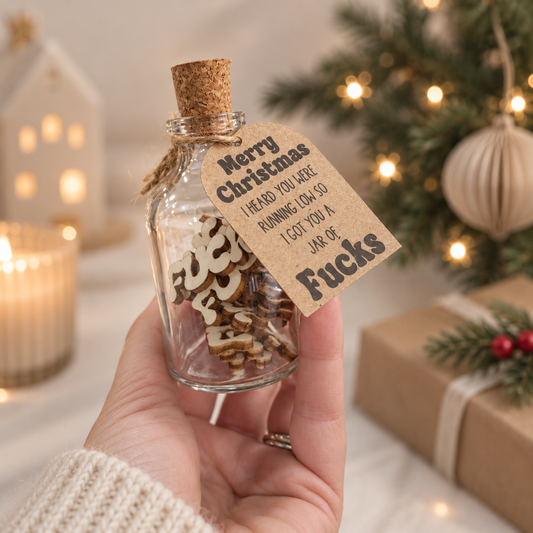 A hand holds a small clear glass bottle with a cork stopper and a hanging kraft paper tag that reads, "Merry Christmas I HEARD YOU WERE RUNNING LOW SO I GOT YOU A JAR OF Fucks." The bottle contains wooden letter cutouts spelling FUCK.