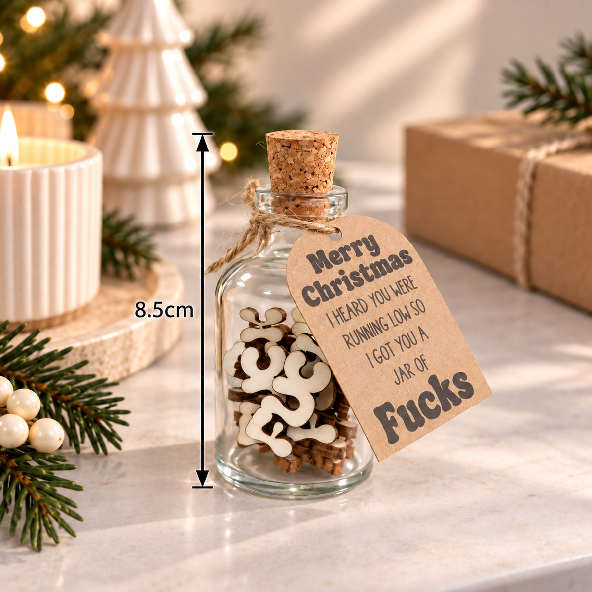 A small clear glass bottle with a cork stopper and a hanging kraft paper tag, filled with wooden novelty letter pieces. The bottle sits on a marble surface with holiday decor, a candle, pine branches, and wrapped gifts in the background.