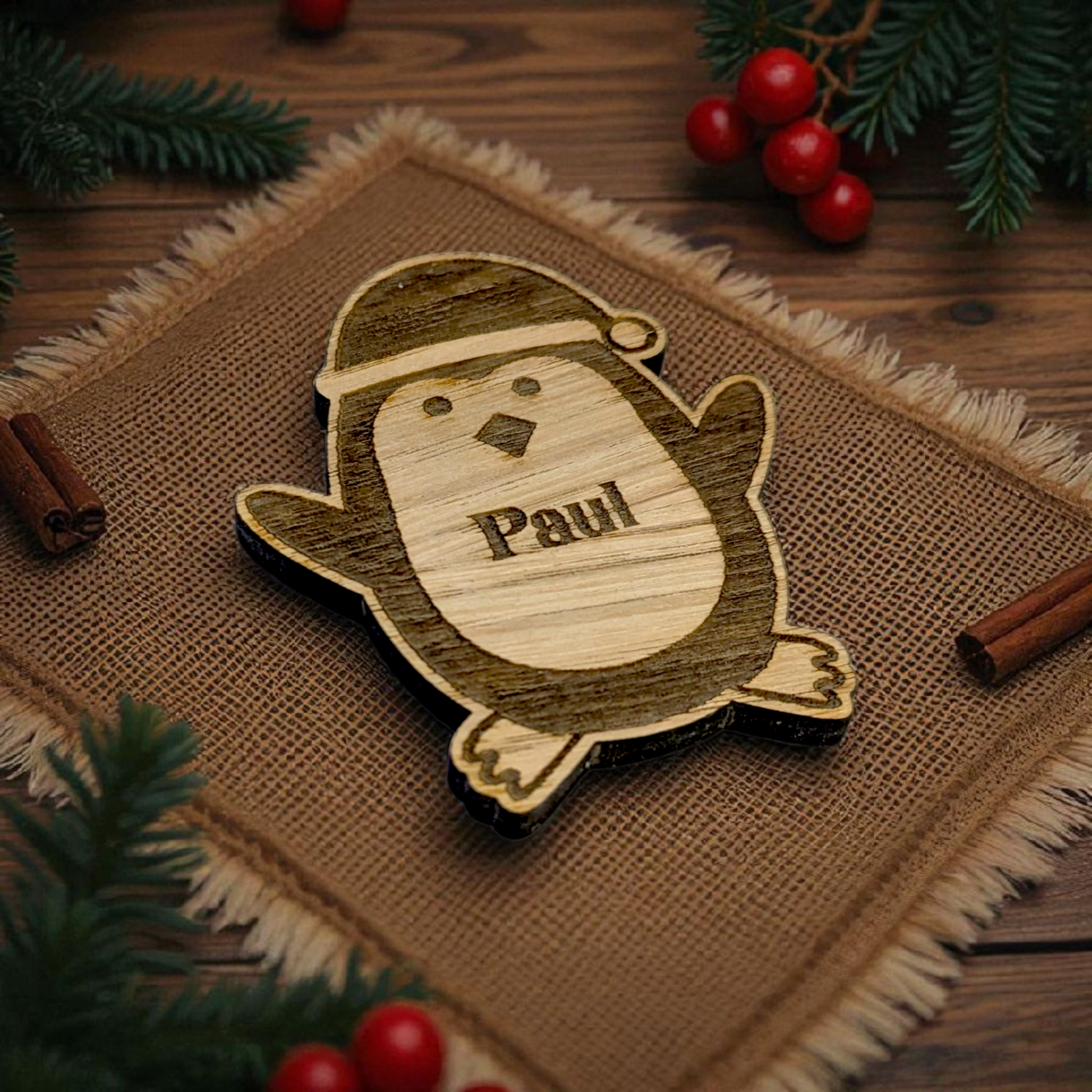 Wooden penguin christmas table gift with 'Paul' engraved, on a rustic background with cinnamon sticks and berries.