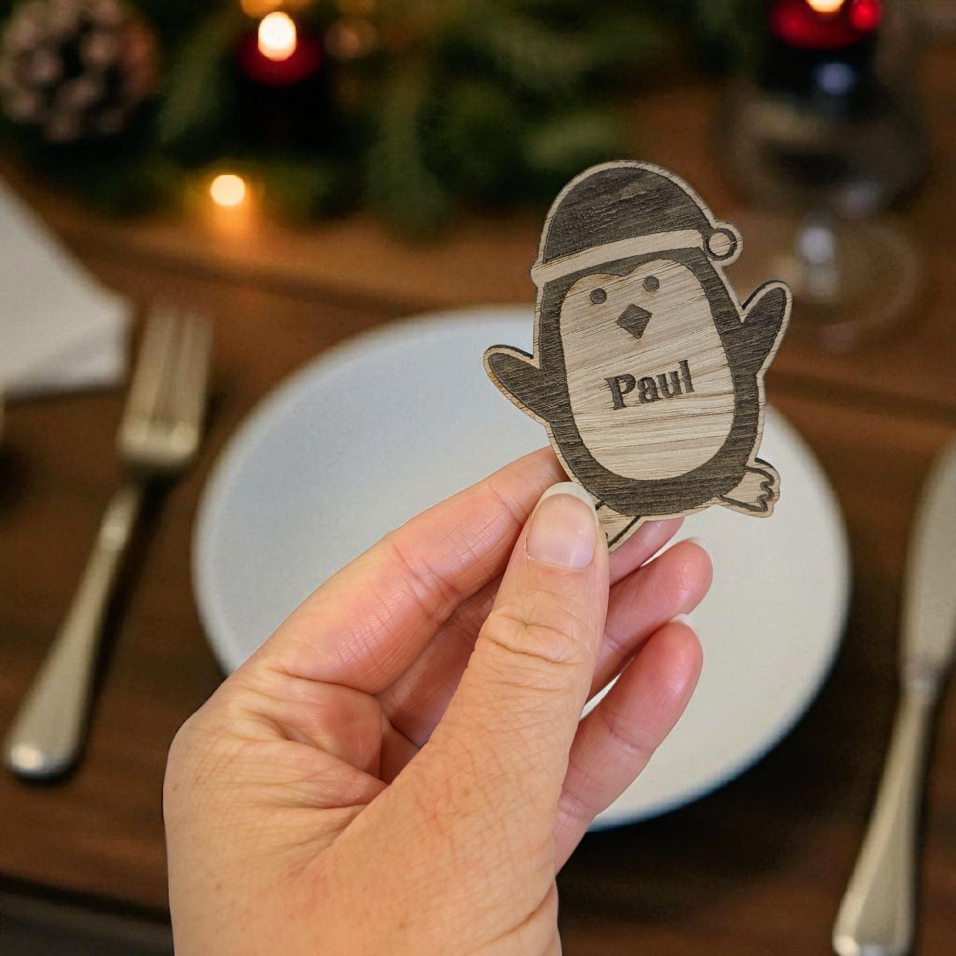 Hand holding a wooden penguin name tag with 'Paul' on a Christmas-themed table.