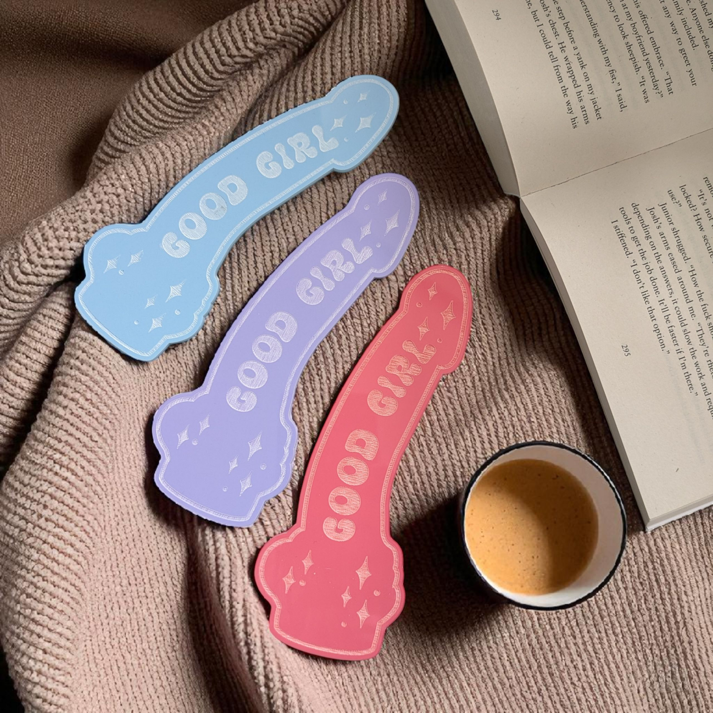 Three 'GOOD GIRL' bookmarks in blue, purple, and pink on a textured surface with a cup and book.