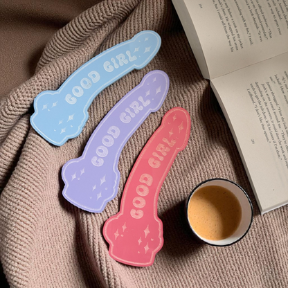 Three 'GOOD GIRL' bookmarks in blue, purple, and pink on a textured surface with a cup and book.