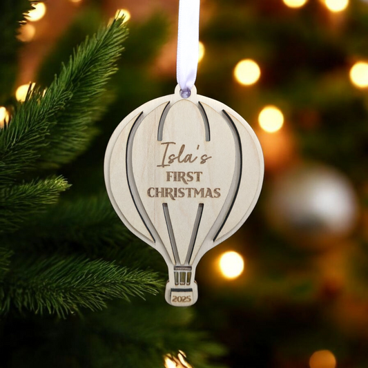 Wooden hot air balloon bauble 'Isla's First Christmas 2025' text on a Christmas tree.