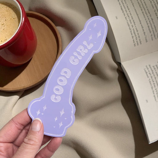 Hand holding a purple 'GOOD GIRL' dildo shaped bookmark with a cup and book in the background.
