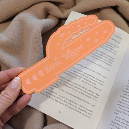 Orange bookmark with 'Book Whore' text held over an open book.