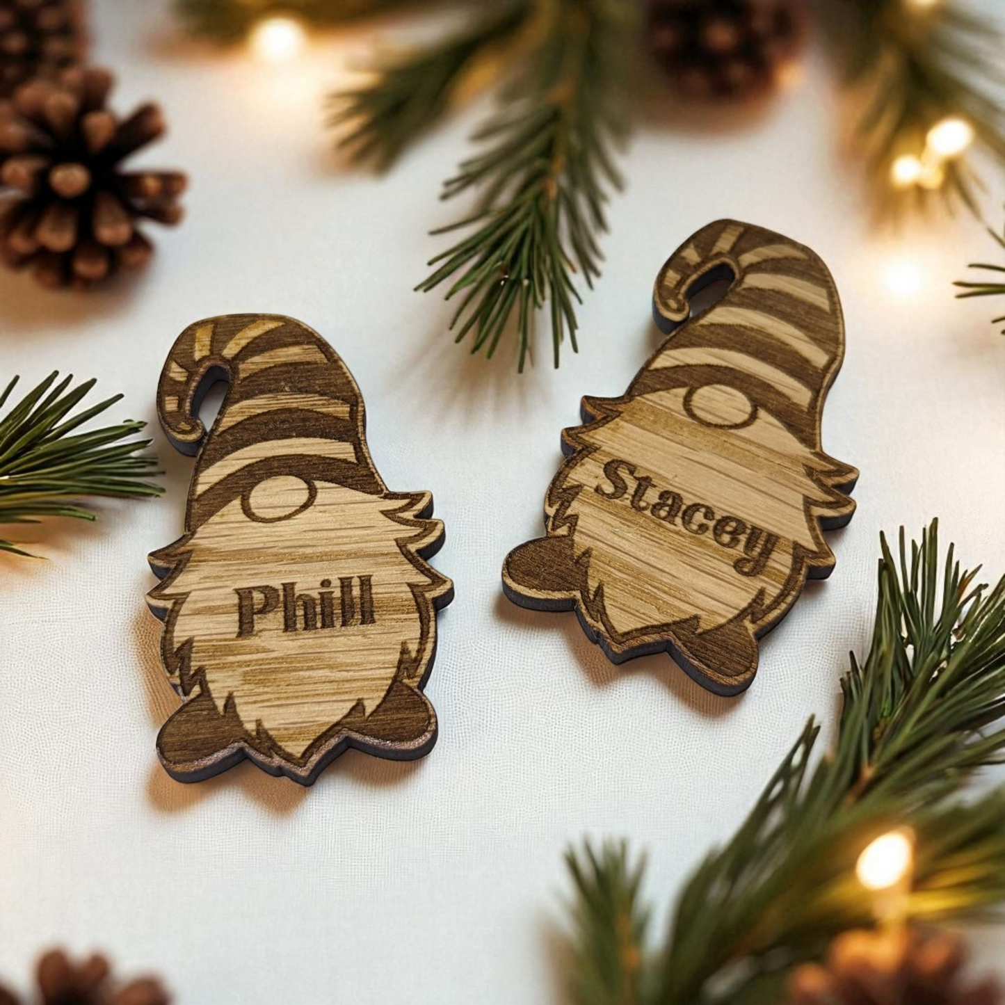 Two christmas wooden gnome table favours with engraved names on a festive background with pine branches and lights.
