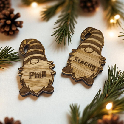 Two christmas wooden gnome table favours with engraved names on a festive background with pine branches and lights.