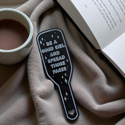 Black spanking paddle bookmark with be a good girl quote on a textured surface with a cup and book.