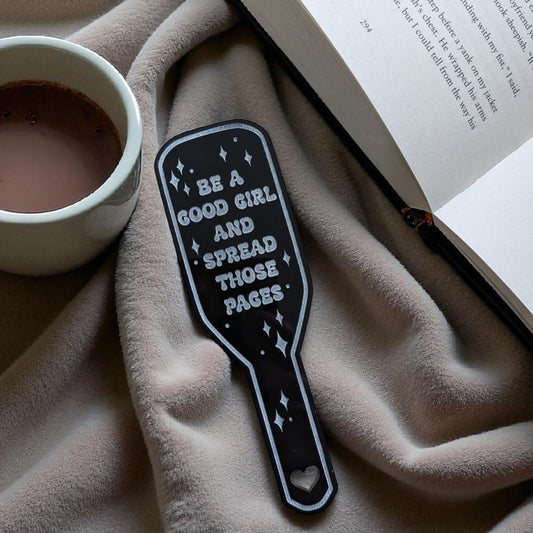 Black spanking paddle bookmark with be a good girl quote on a textured surface with a cup and book.