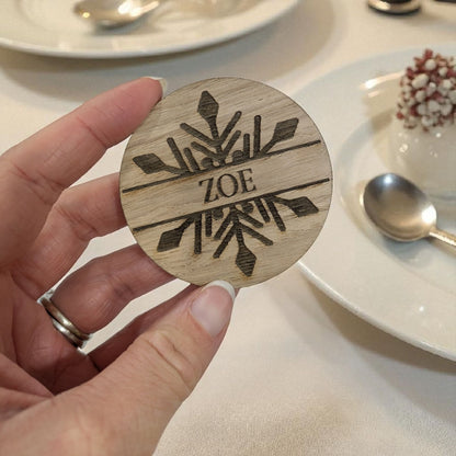 Wooden christmas table favour with engraved design and 'ZOE' text held by a hand, with a dining setting in the background.