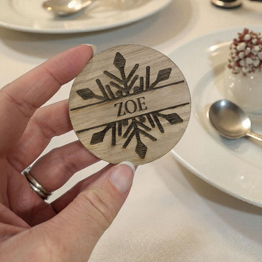 Wooden christmas table favour with engraved design and 'ZOE' text held by a hand, with a dining setting in the background.