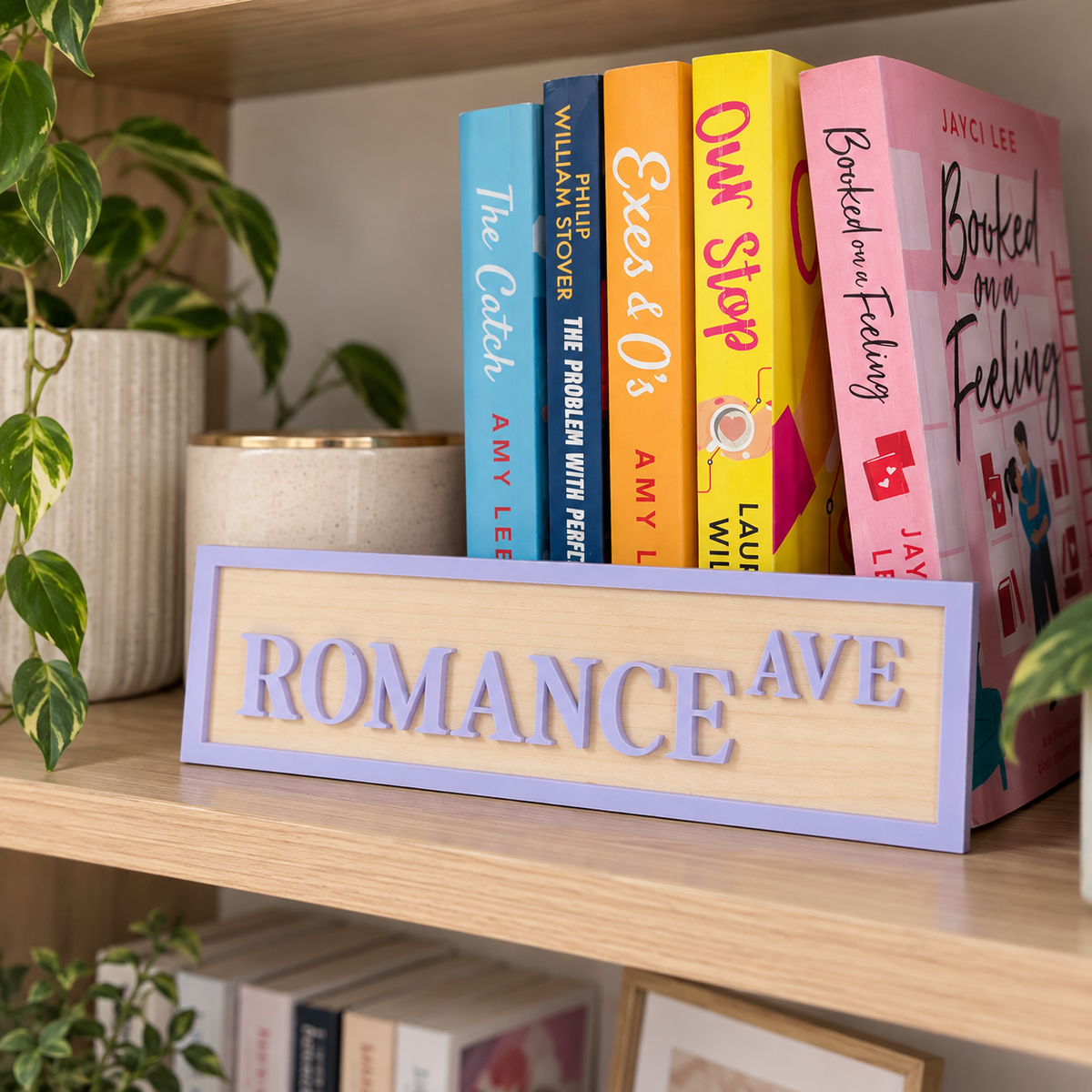 A rectangular wooden sign with raised letters spelling 'ROMANCE AVE' in lavender, displayed on a wooden bookshelf next to books and potted plants.