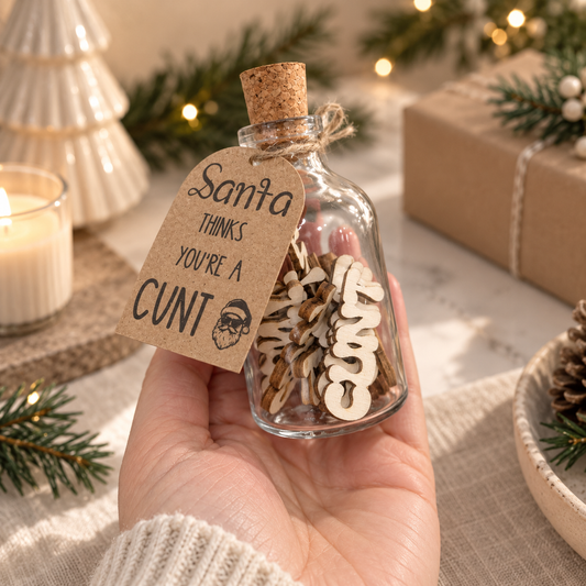 A hand holds a small clear glass bottle with a cork stopper, filled with wooden Christmas letter decorations and a kraft paper tag that reads, 'Santa thinks YOU'RE A CUNT.' The scene is styled with holiday greenery, a candle, and wrapped gifts in the background.