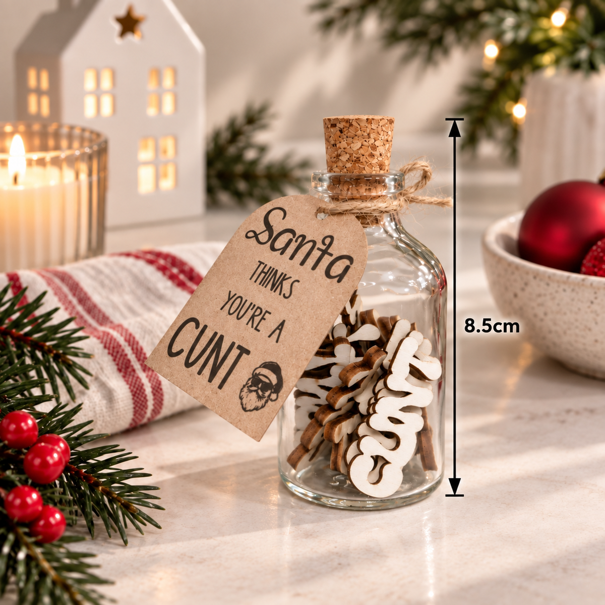 A small clear glass bottle with a cork stopper and a hanging kraft paper tag reading 'Santa THINKS YOU'RE A CUNT' is styled on a marble surface with holiday decor, a candle, pine branches, and red ornaments. A vertical measurement marker on the right shows 8.