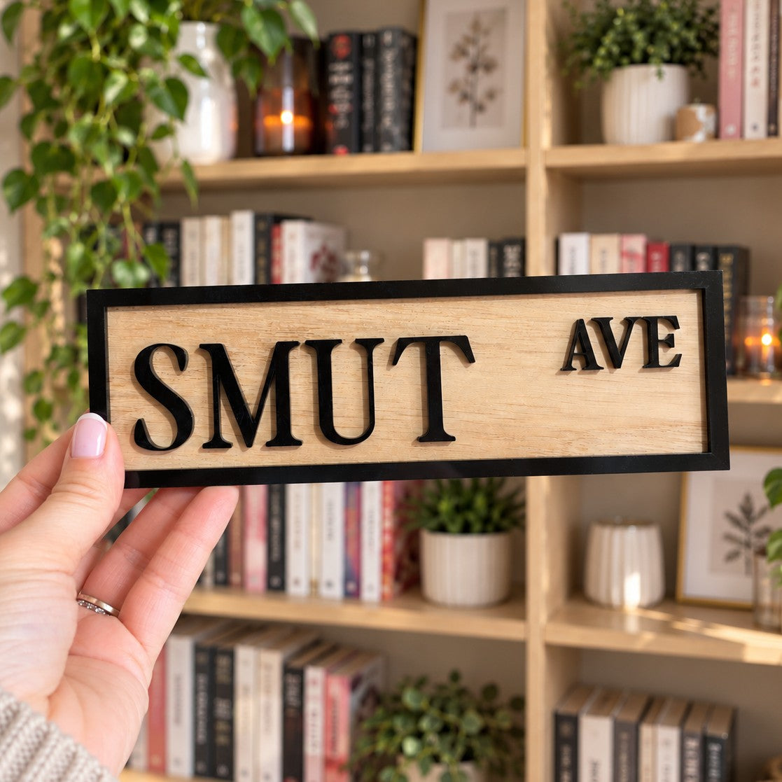 A hand holds a rectangular wooden sign with the words 'SMUT AVE' in black serif letters on a light natural wood background with a black border. The background shows a softly blurred bookshelf with books and plants.