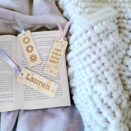 a set of three small bookmarks for adults. One reads hot girls read smut, another with 3 different daisy designs and the last with a cute floral design and a name. All details are engraved in to the wooden bookmarks.