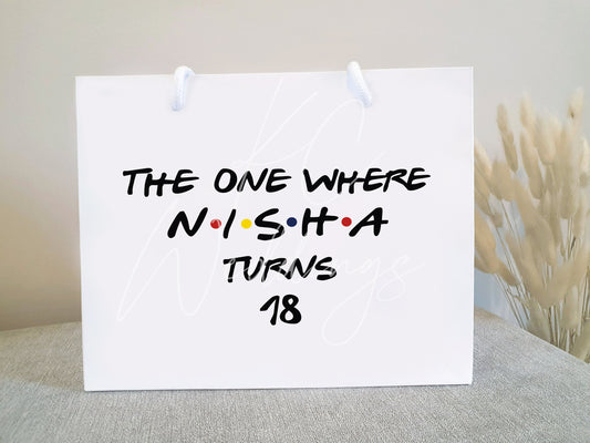 a small white birthday gift bag that says ' the one where Nisha turns 18' made with the friends tv show in mind.